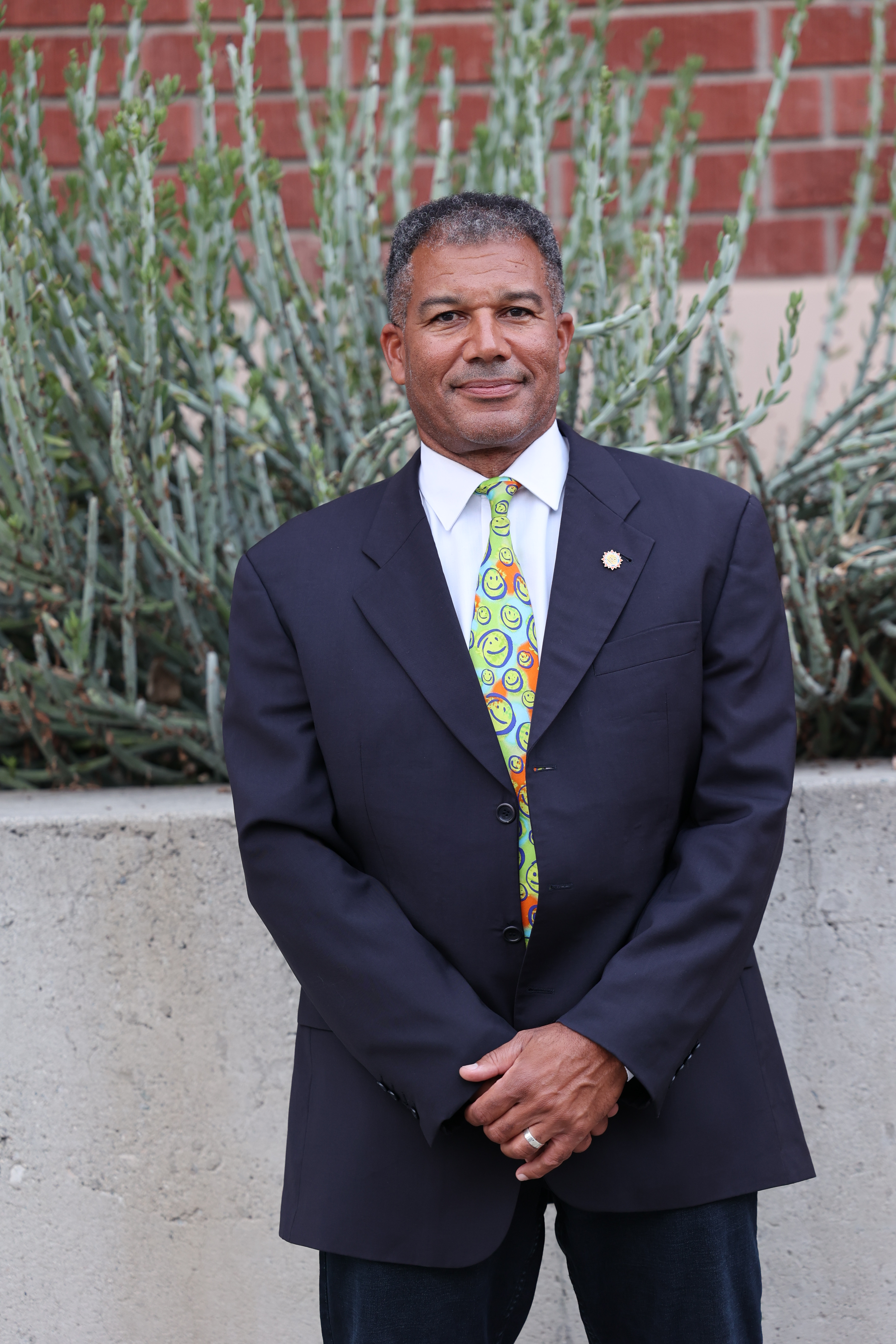 Ray Jackson, wearing a navy suit and patterned tie, standing outdoors in front of shrubs and a brick wall.