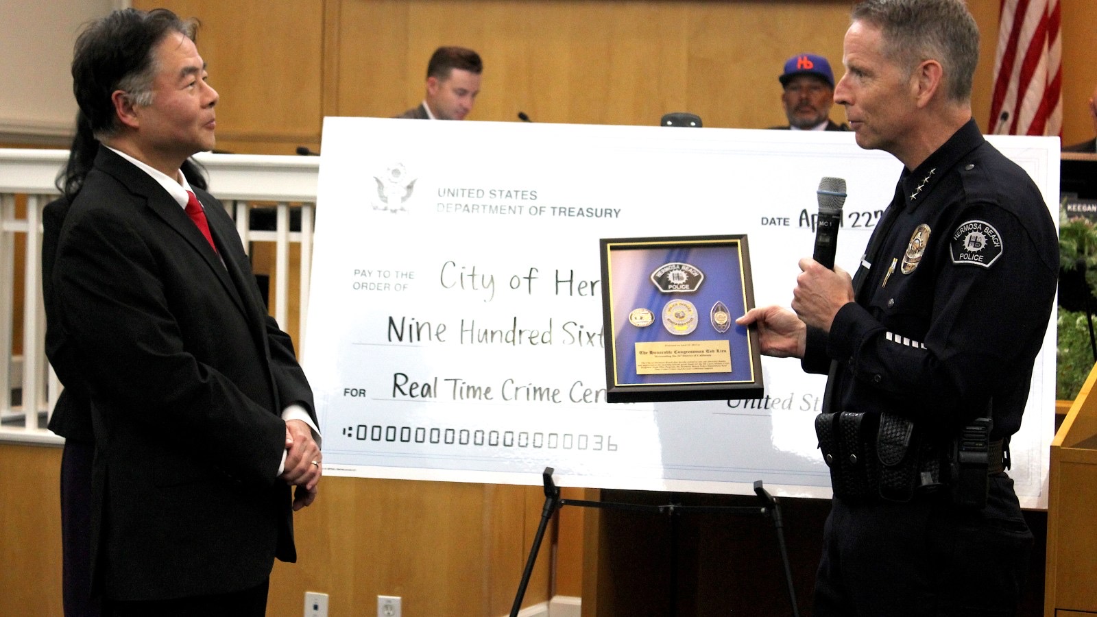 A police officer presenting a plaque to another man at a podium, with a U.S. Treasury check displayed nearby.